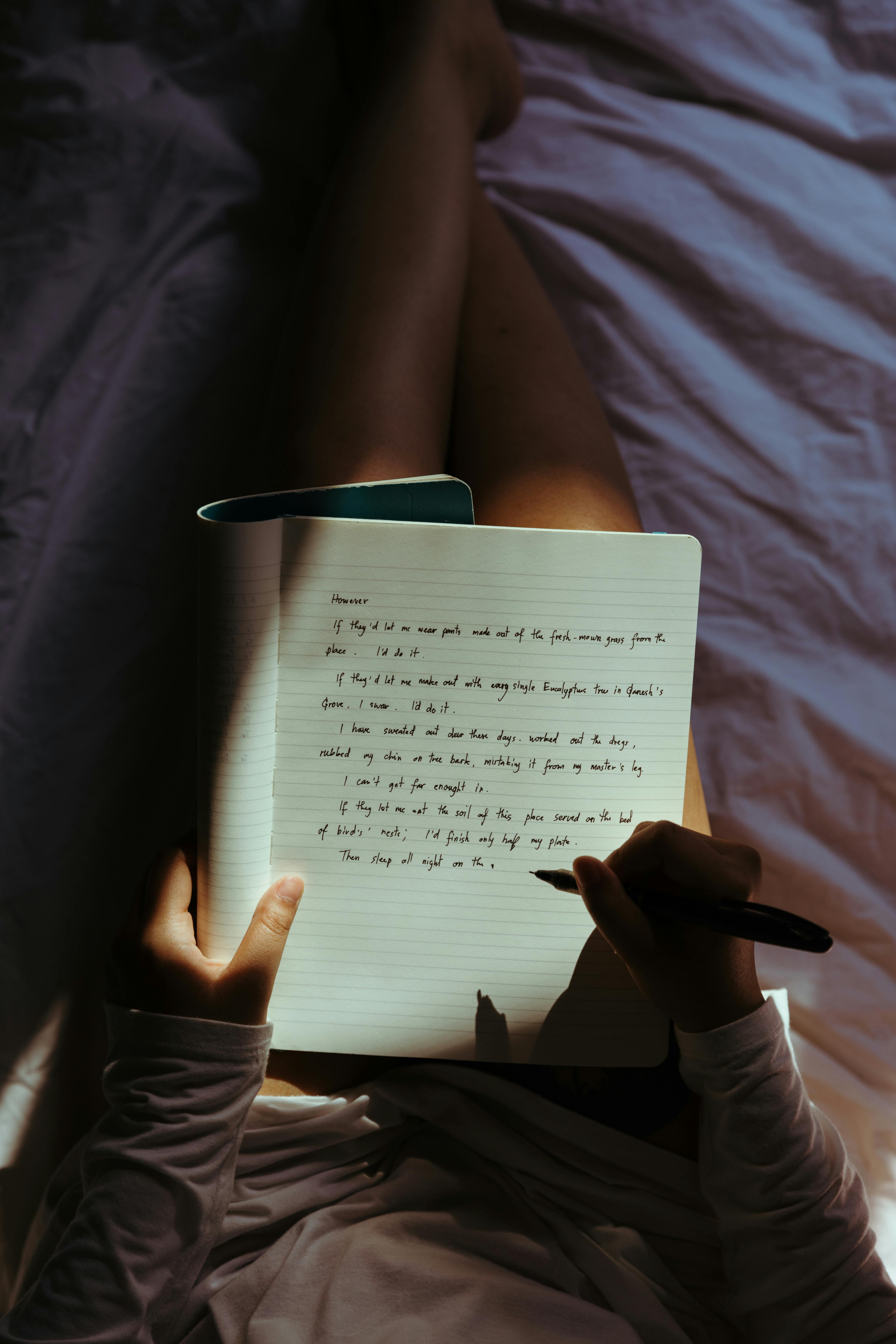 Woman writing in a notebook on her lap while relaxing comfortably in a cozy, dimly lit bedroom setting.