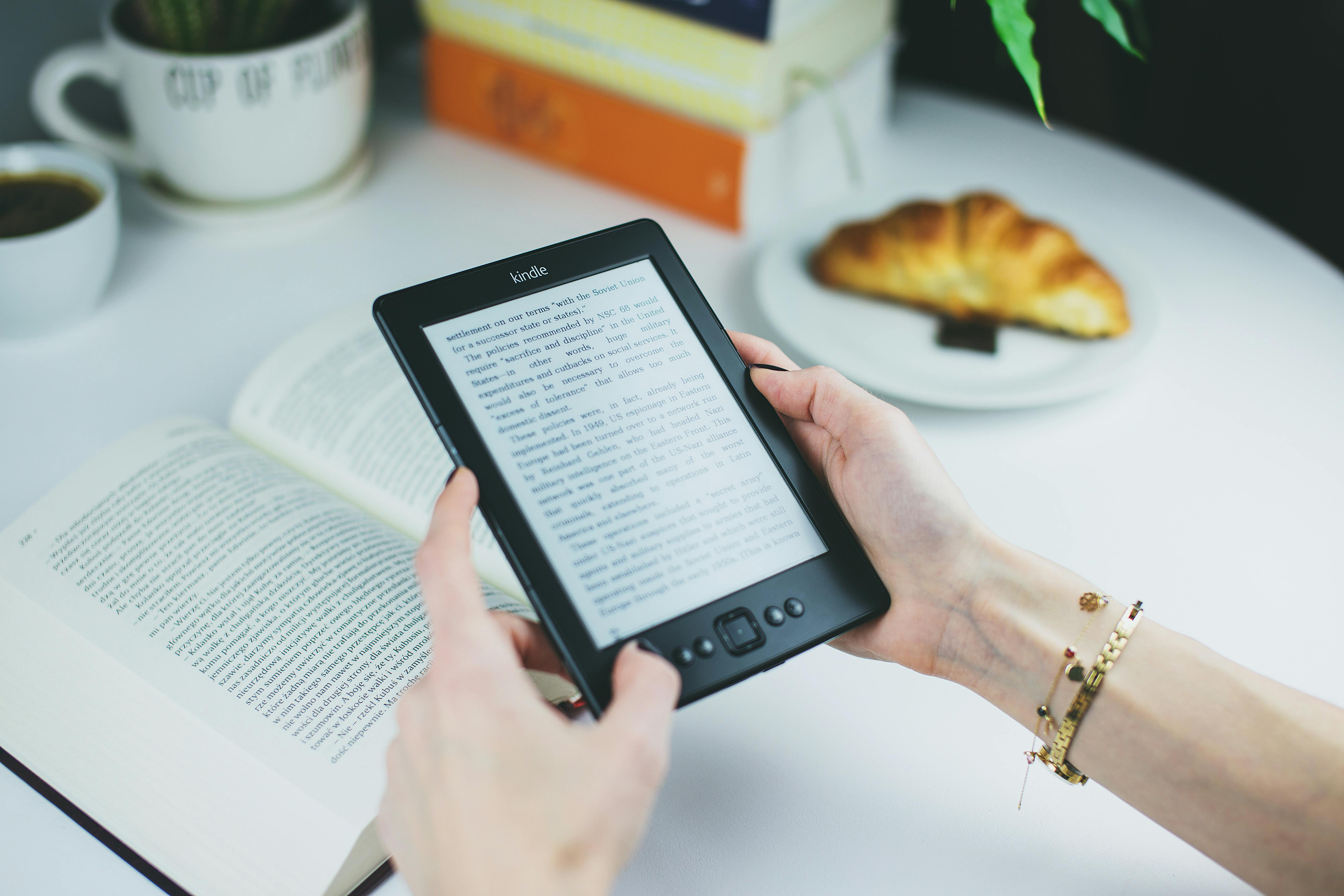 A woman holds a Kindle while reading with a book and croissant on a table.